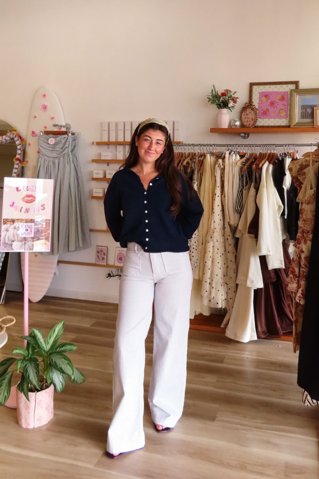 A woman stands in a boutique wearing a navy blue long-sleeve button-up top and white wide-leg pants. The background shows clothing racks with various garments, a wooden floor, and a beige wall.