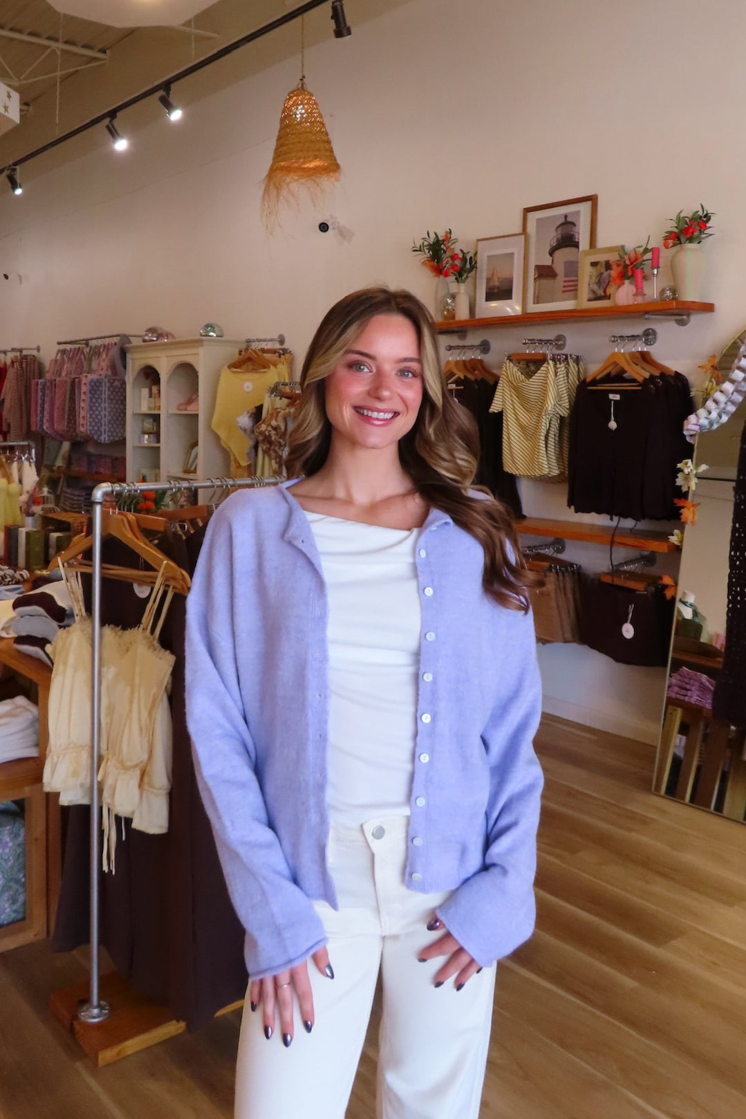 A woman stands in a boutique wearing a light lavender cardigan over a white top and white pants. The background shows clothing racks, shelves, and store fixtures.