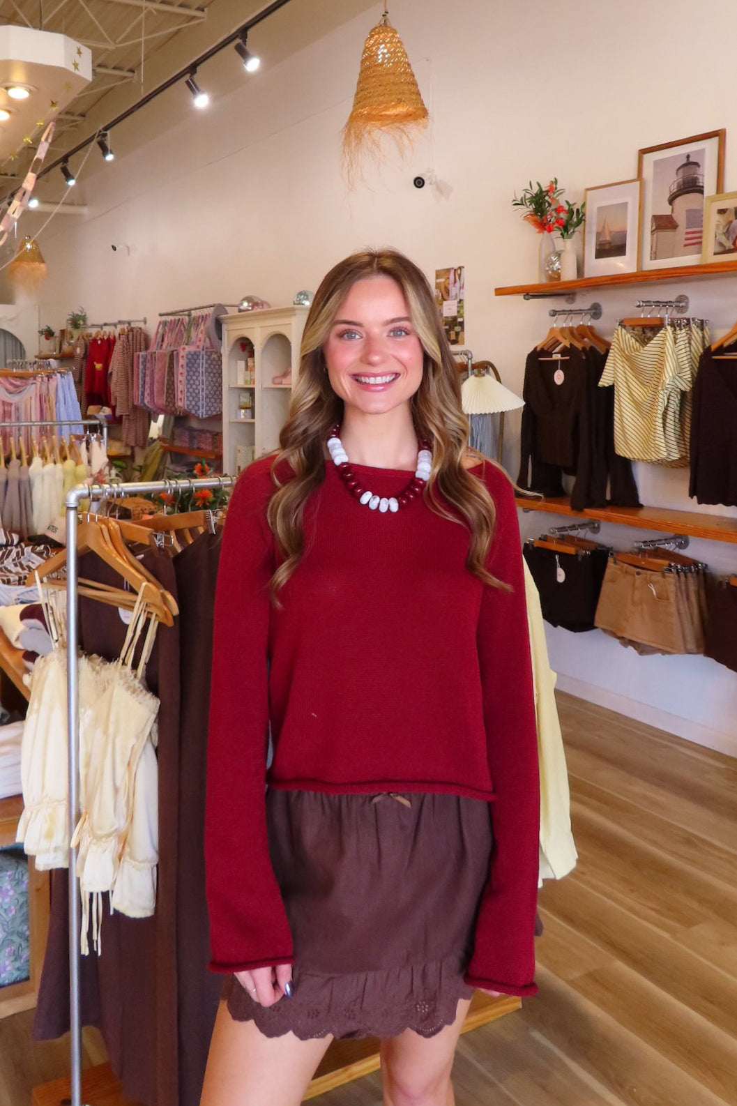 A smiling woman stands inside a boutique wearing a long-sleeve burgundy knit sweater and a brown ruffled skirt, accessorized with a chunky white beaded necklace.