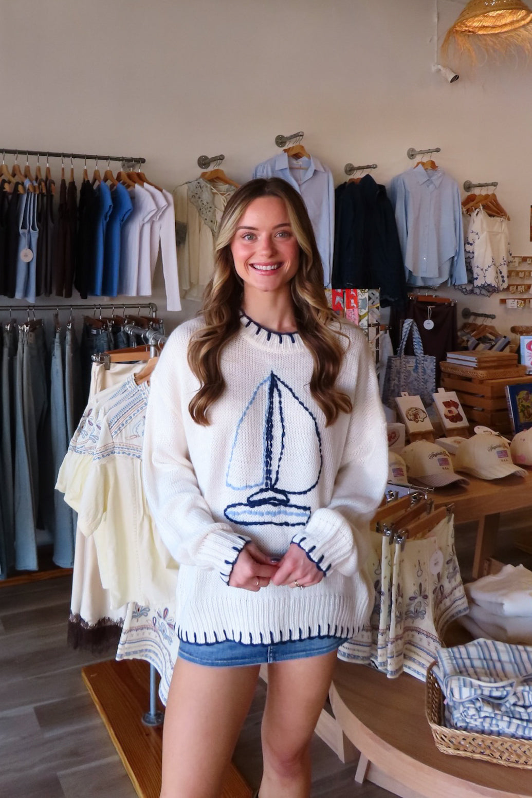 A woman stands inside a clothing boutique wearing a cream knit sweater with a navy blue sailboat graphic on the front, paired with denim shorts. The background shows racks of shirts, dresses, and other apparel items.