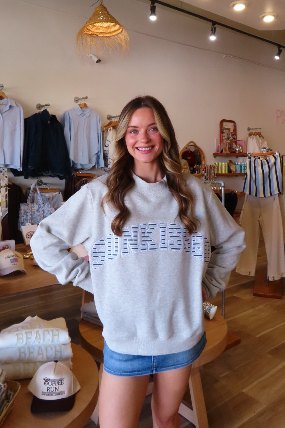 A woman smiling in a boutique while wearing a light gray sweatshirt with a blue graphic text print across the chest, paired with a denim mini skirt. The store background features clothing racks and wooden floors.