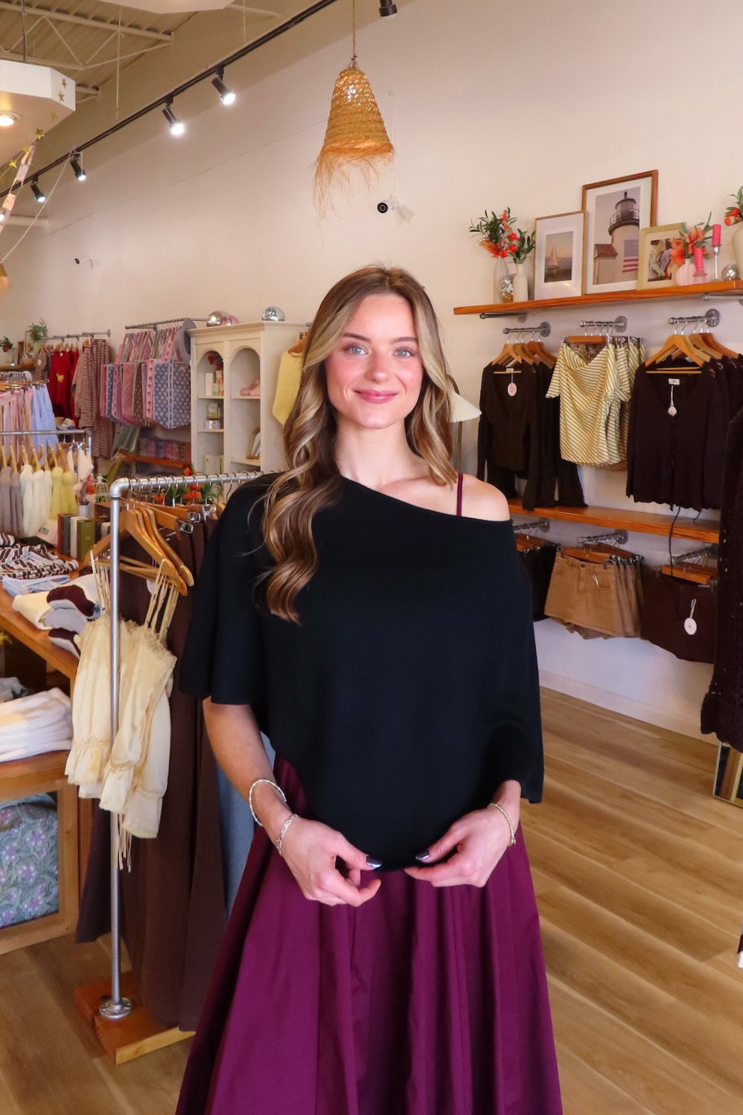A woman standing inside a clothing boutique wearing a black off-the-shoulder top and a dark purple skirt, with racks of garments and wooden floors visible in the background.