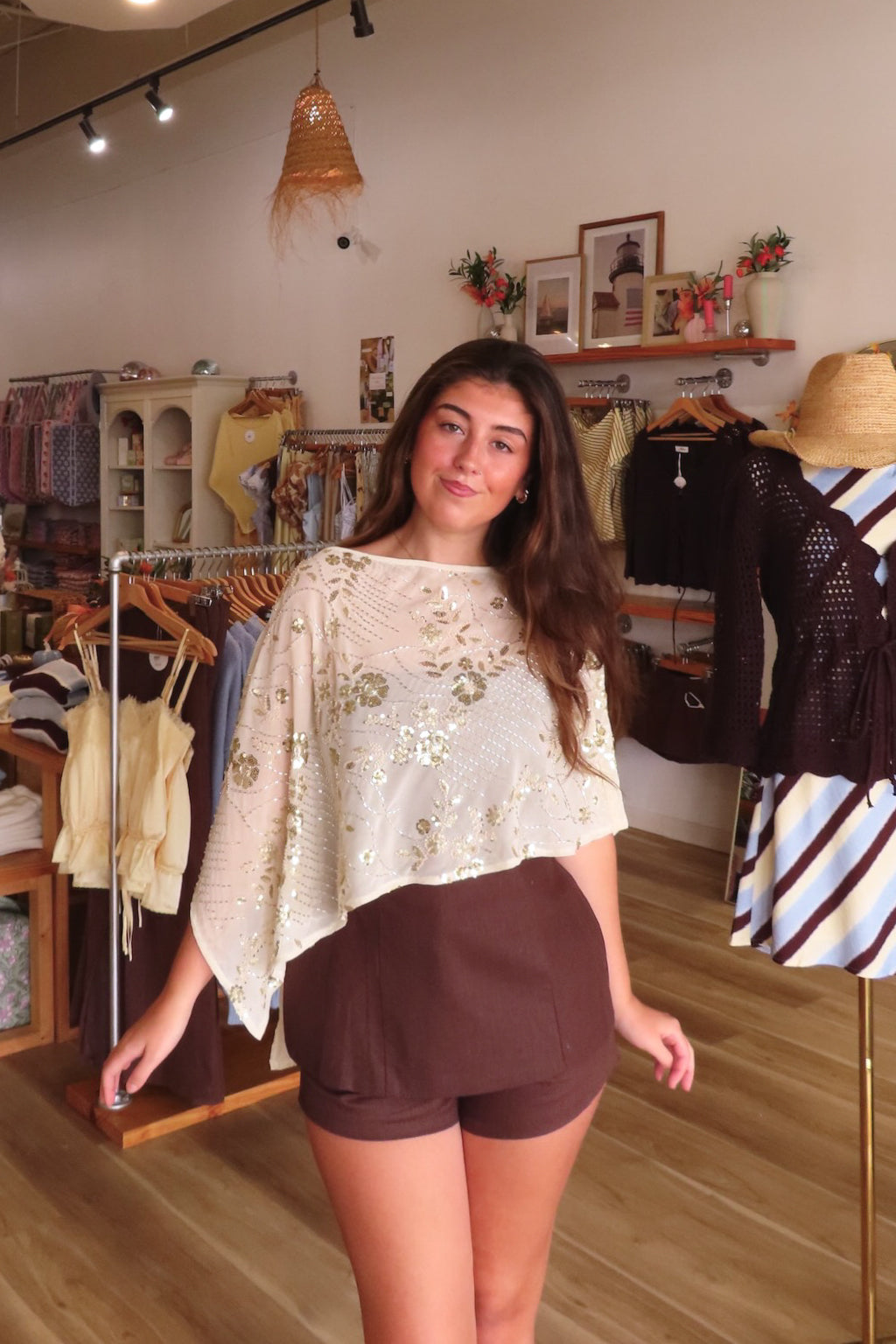 A woman modeling a cream poncho top with gold sequin embroidery and brown shorts inside a boutique clothing store.