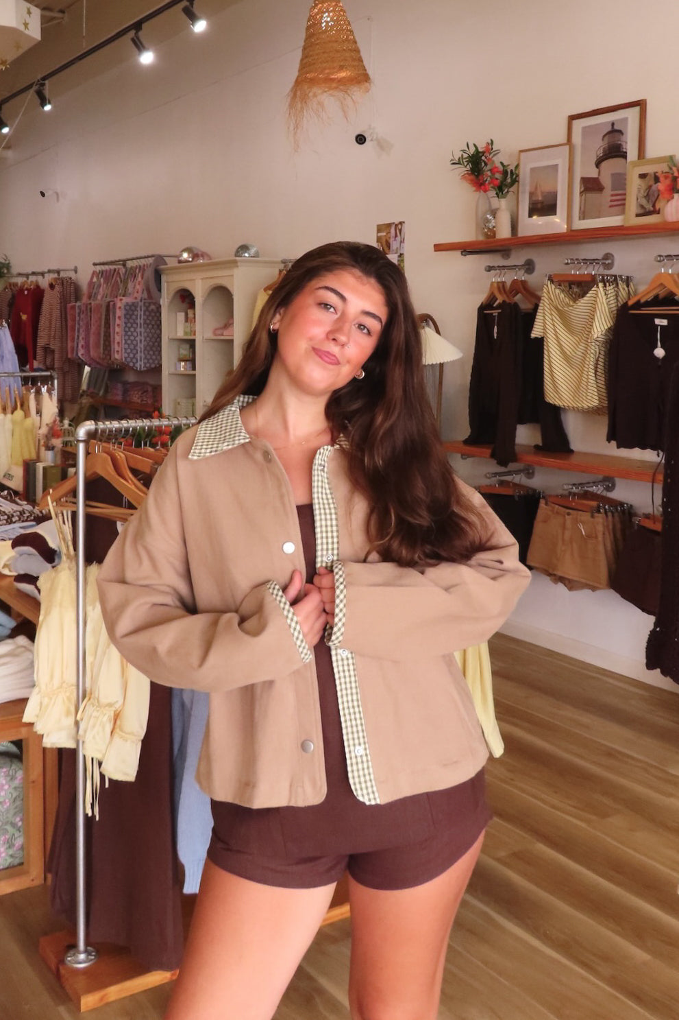 A woman modeling a beige cropped button-up shirt jacket with a green and white gingham patterned collar and placket, paired with brown shorts inside a boutique clothing store.