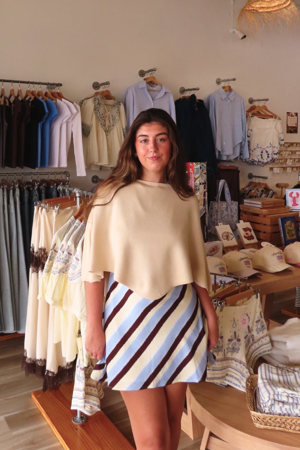 A woman modeling a beige cape-style poncho top paired with a blue, white, and brown striped mini skirt inside a boutique clothing store.