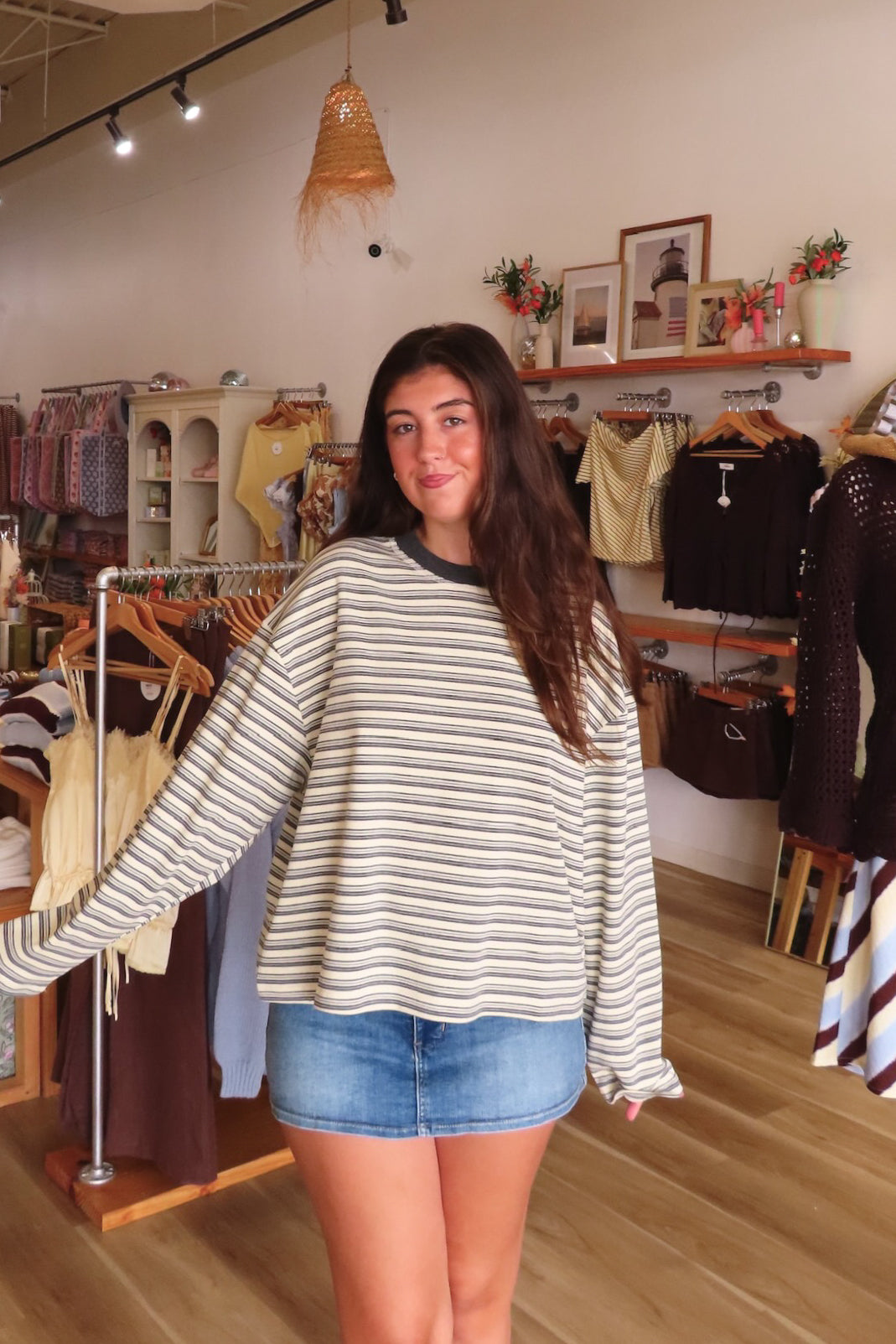 A woman modeling a cream and dark gray striped long-sleeve knit sweater with a crew neck, paired with denim shorts inside a boutique clothing store.