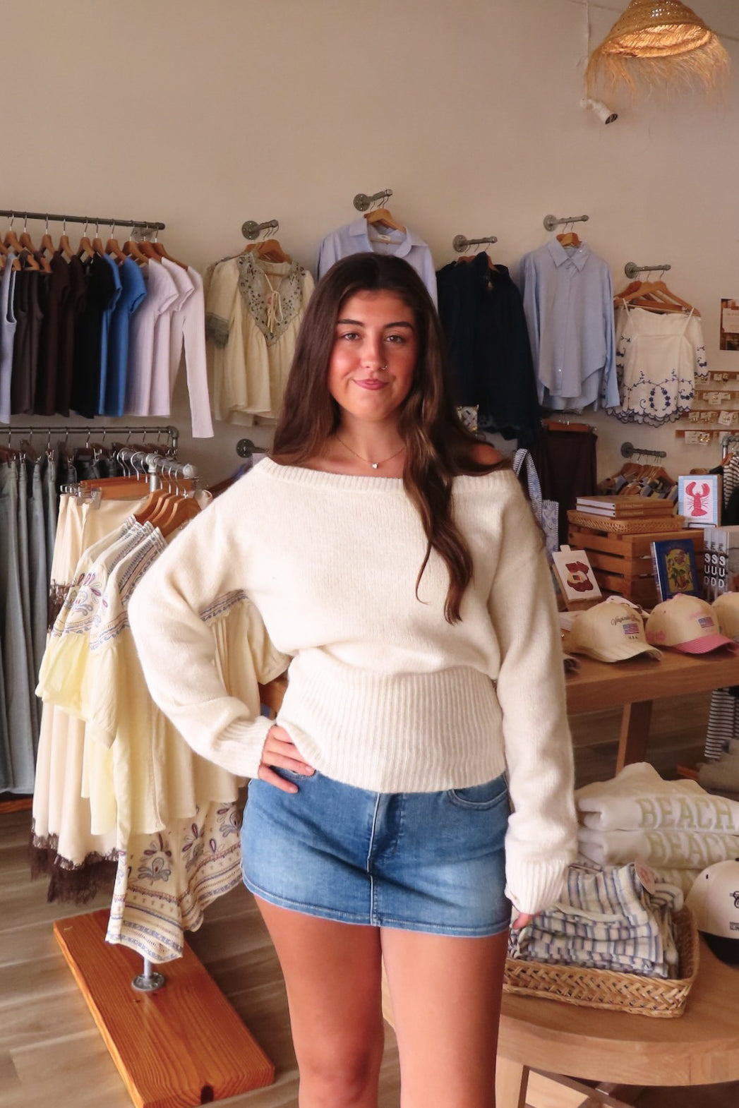A woman modeling a cream off-the-shoulder knit sweater and denim shorts inside a boutique clothing store with racks of garments and wooden display tables in the background.